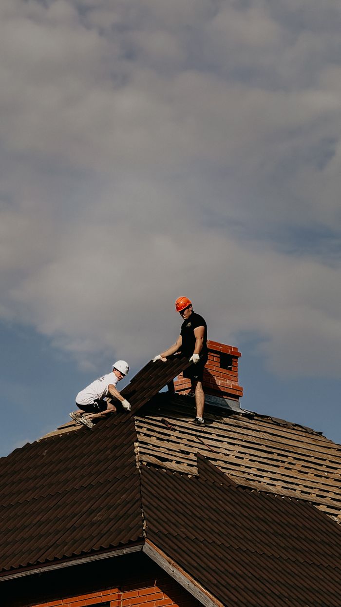 hero-img-02 Two workers in safety helmets repair a roof, showcasing outdoor construction during a cloudy day.